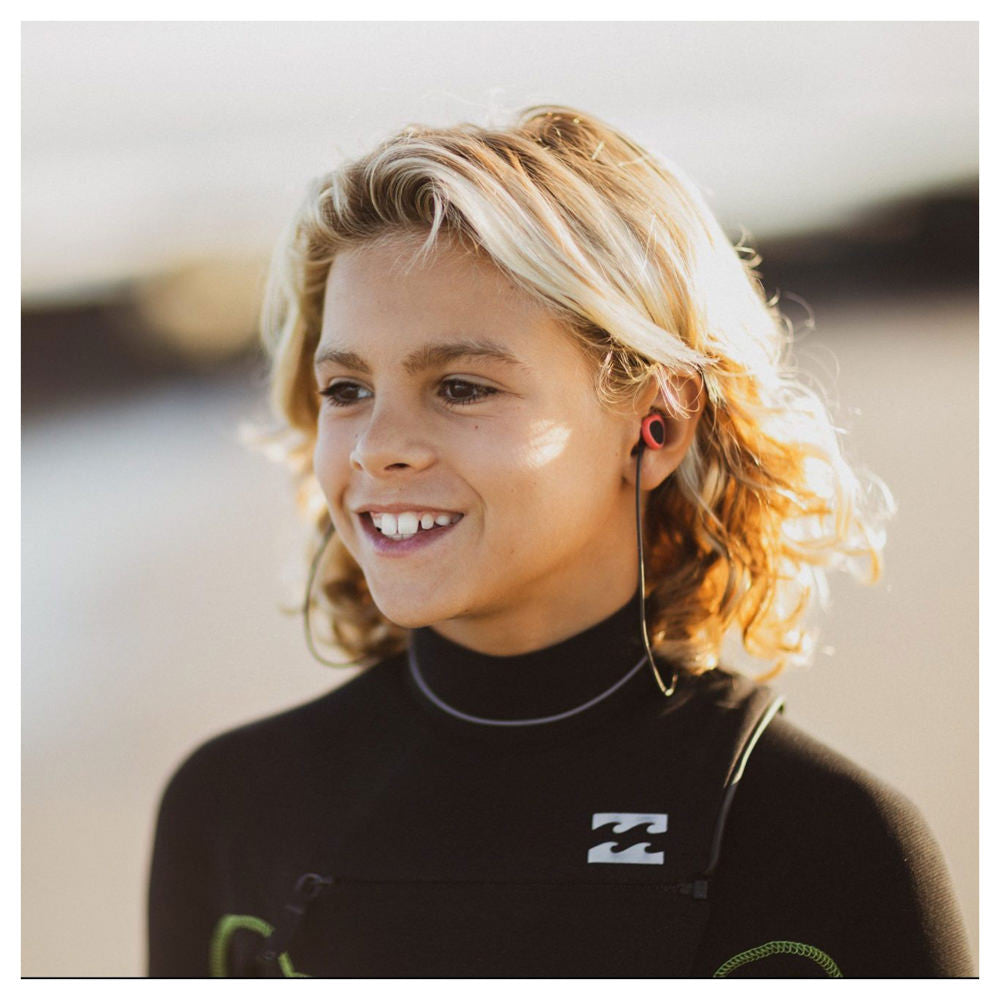 Blondes Kind mit schulterlangen Haaren trägt einen Neoprenanzug und hat SurfEars Junior im Ohr.